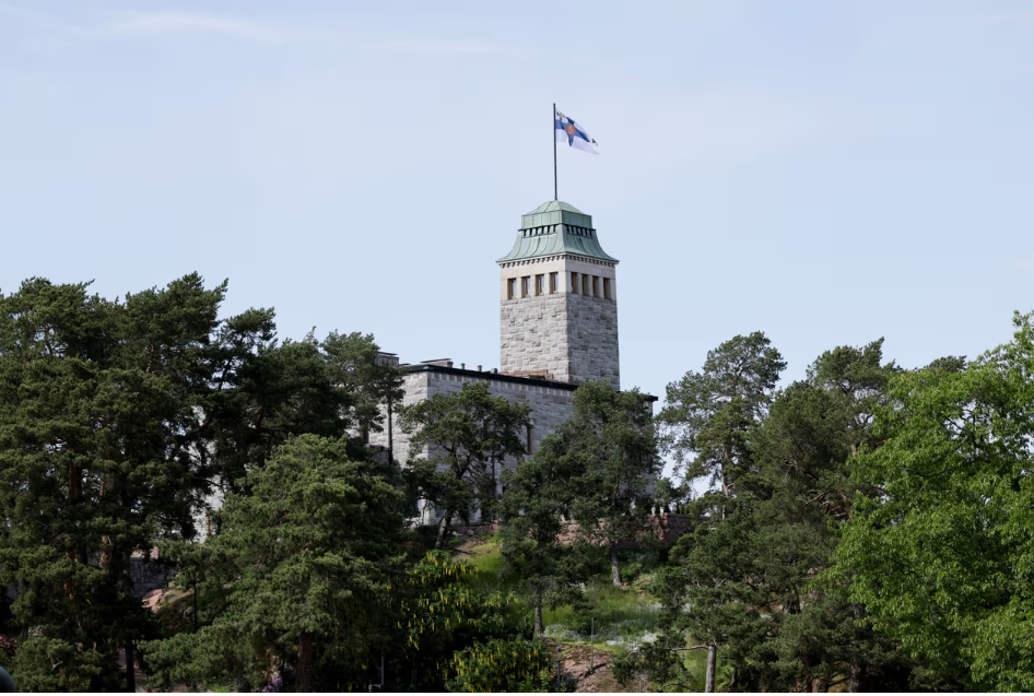 Kuvassa Naantalissa sijaitsevan Kultaranta-rakennuksen kivinen torni, jonka huipulla liehuu tuulessa Suomen lippu. Rakennuksen ympärillä on paljon vihreyttä ja puita. 